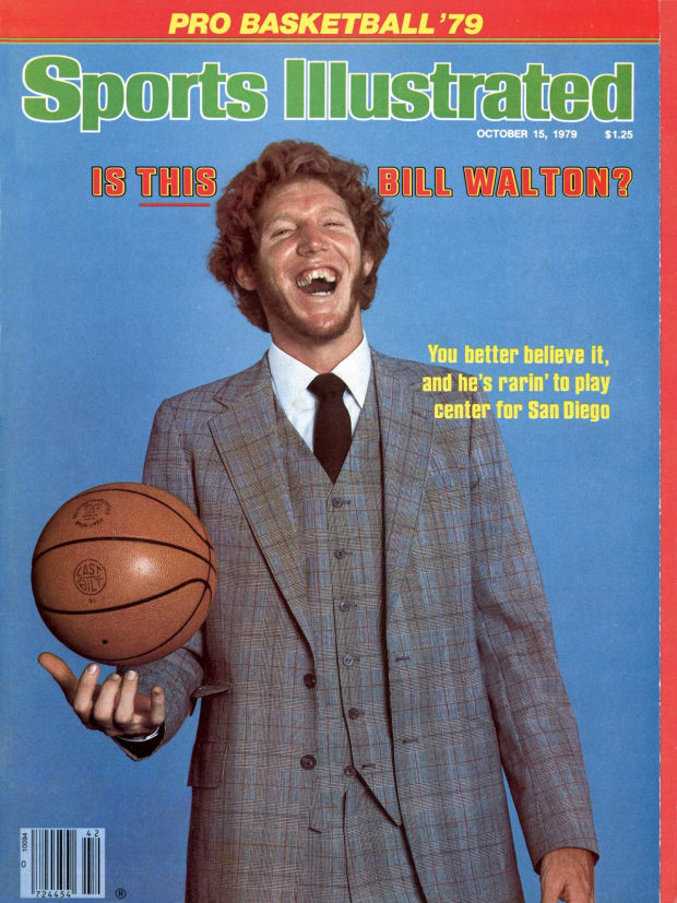 Bill Walton smiles while holding a basketball and wearing as suit. 